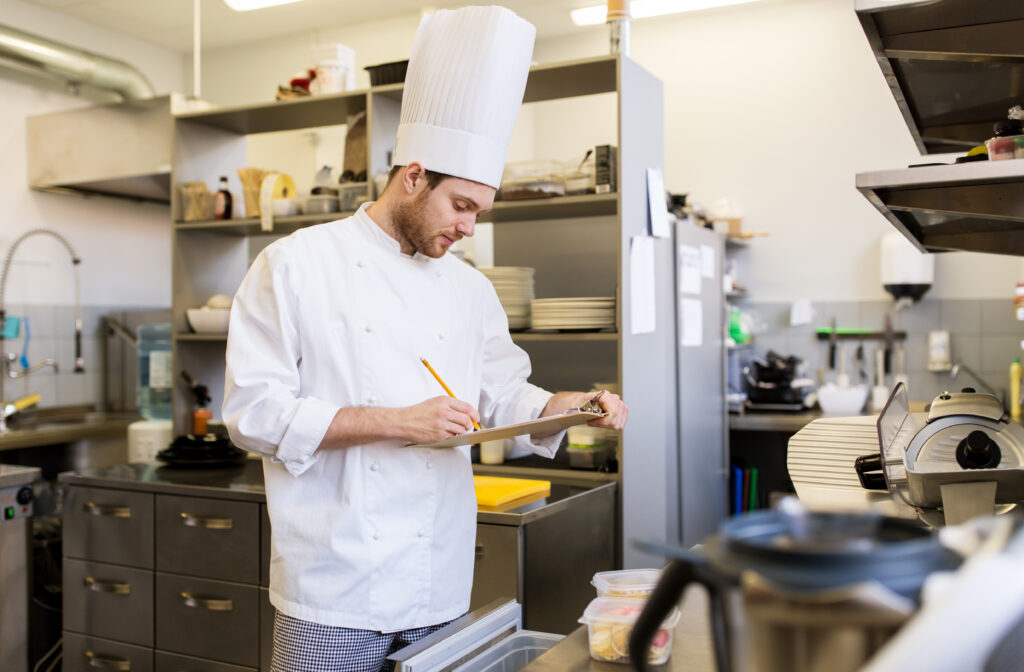 A chef in a hat is taking inventory in a commercial kitchen.