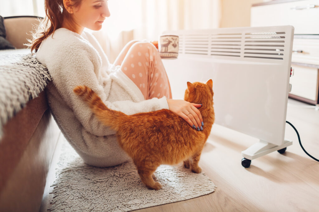 A woman sitting in front of an indoor heater with her cat and holding a cup of tea.