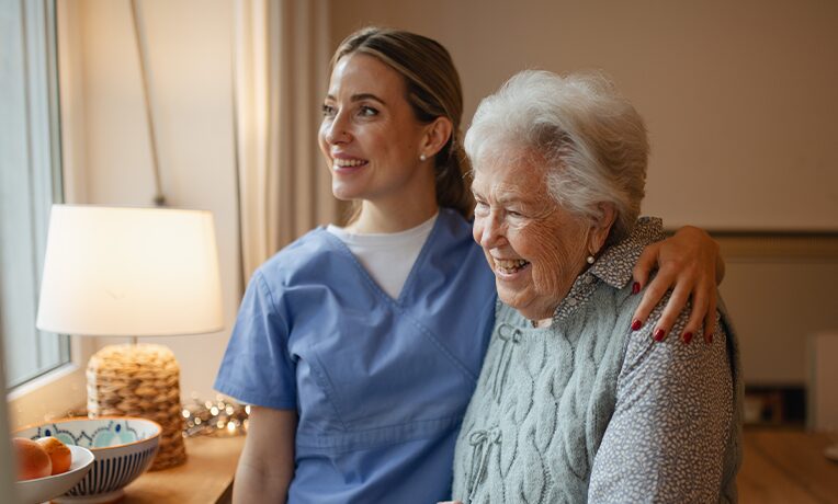 A healthcare worker stands beside an elderly woman, sharing a moment of care and companionship.